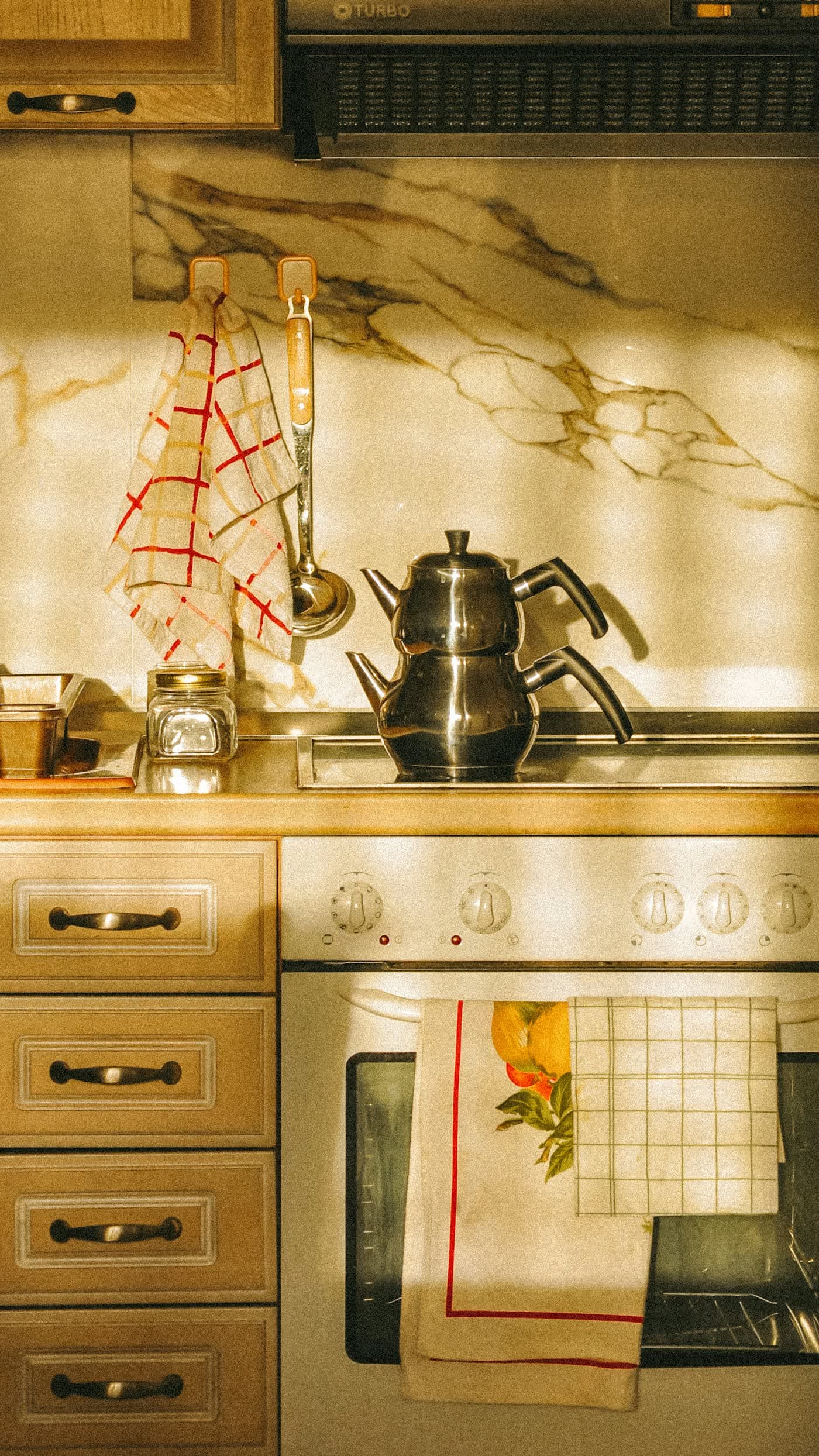 Cozinha pequena sob luz quente da tarde, com pano de prato xadrez, chaleira de inox e fogão antigo