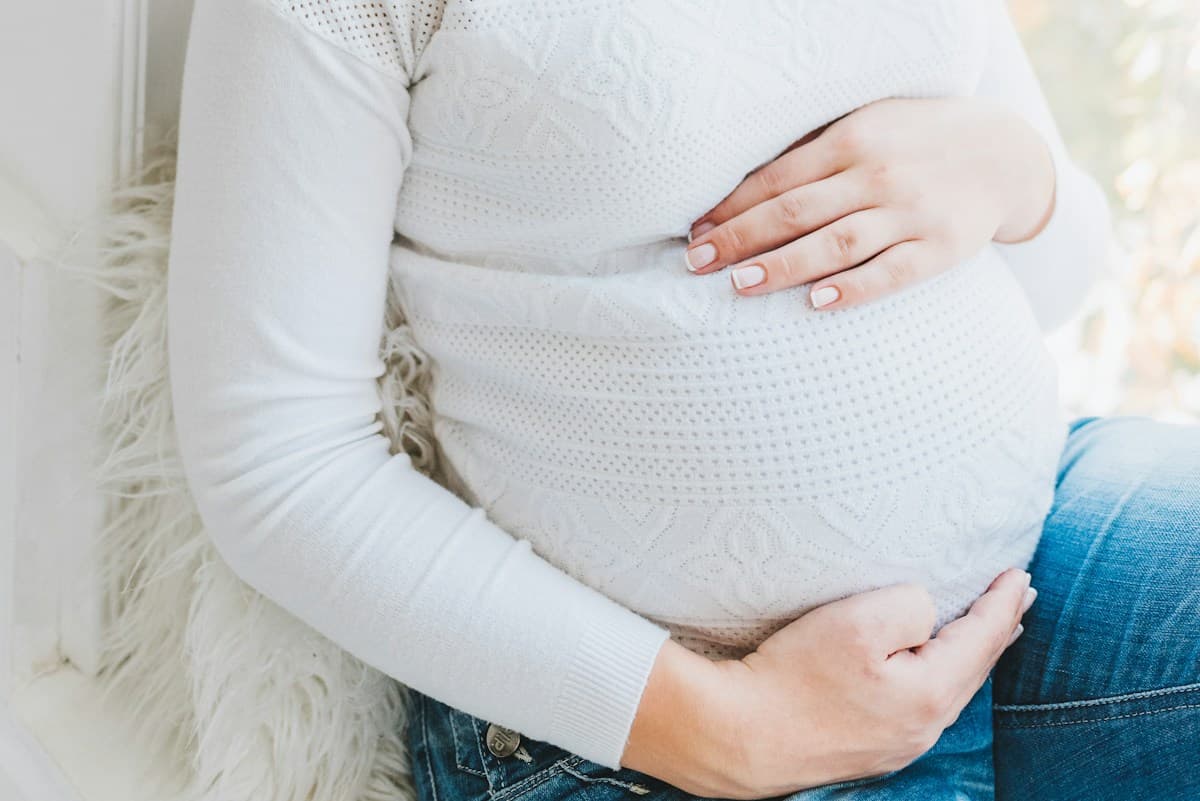 Gestante de suéter de tricô branco com as mãos sobre a barriga, sentada perto da janela