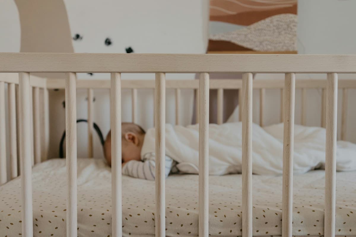 Bebê dormindo de costas em berço de madeira branca com lençol e cobertor claros