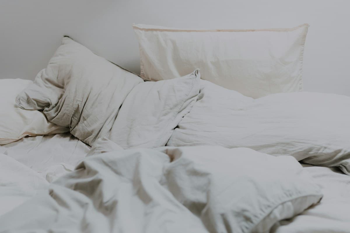Cama desarrumada com travesseiros e roupa de cama branca de algodão sob luz natural