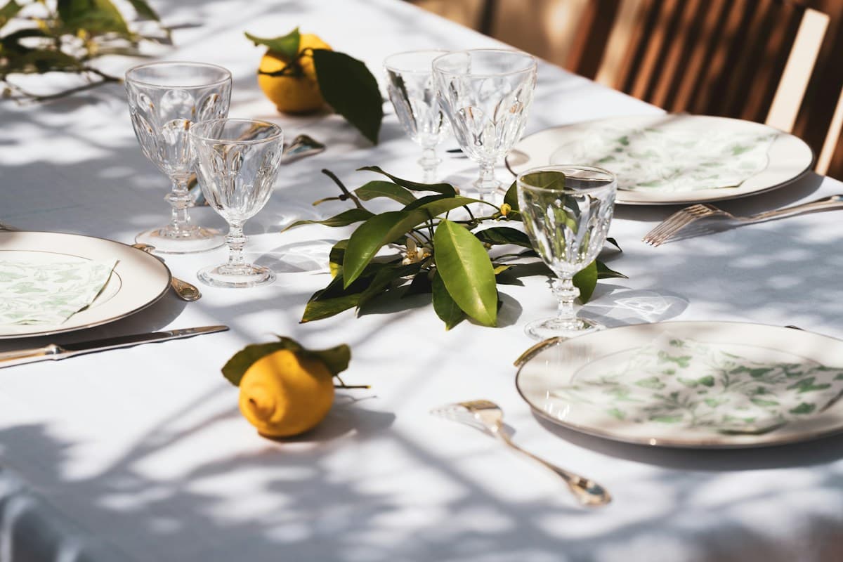 Mesa posta com toalha branca, pratos, taças de cristal, limões e folhas sob sol de tarde