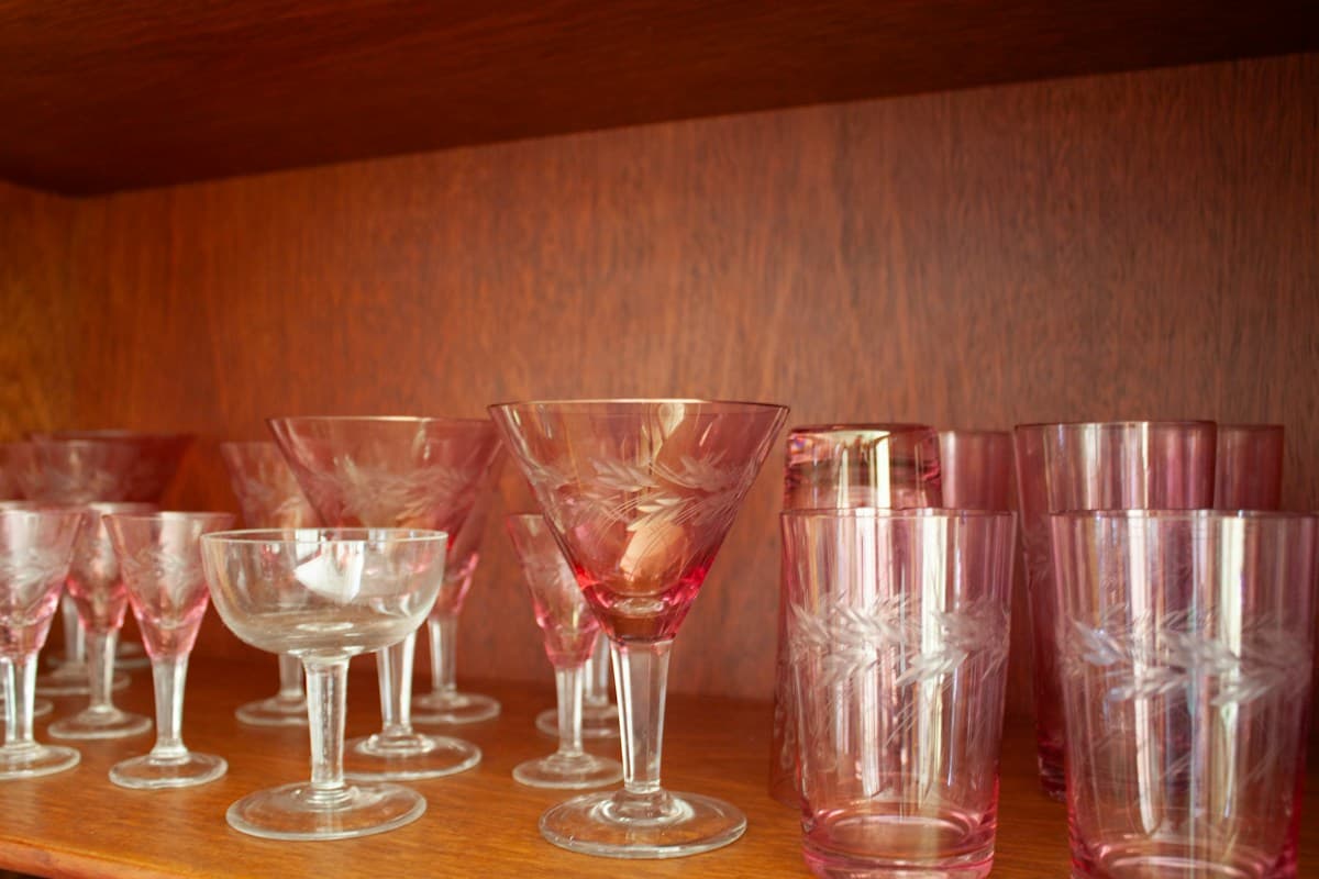 Taças e copos de vidro lapidado, transparentes e rosados, enfileirados em prateleira de madeira