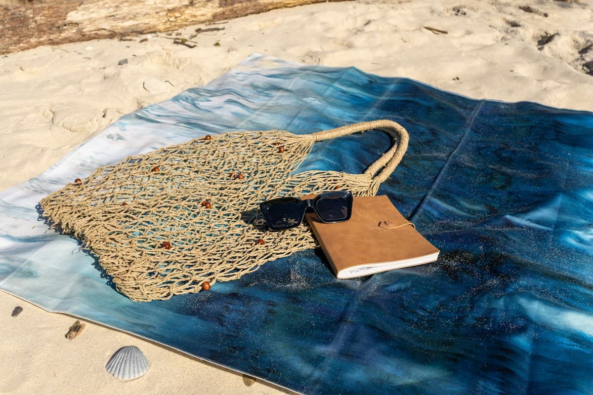 Bolsa de crochê, óculos de sol e caderno sobre canga estendida na areia da praia